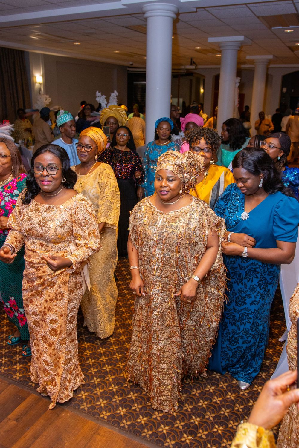 A group of guests in colorful traditional attire dancing together at a formal event with elegant columns and decor.