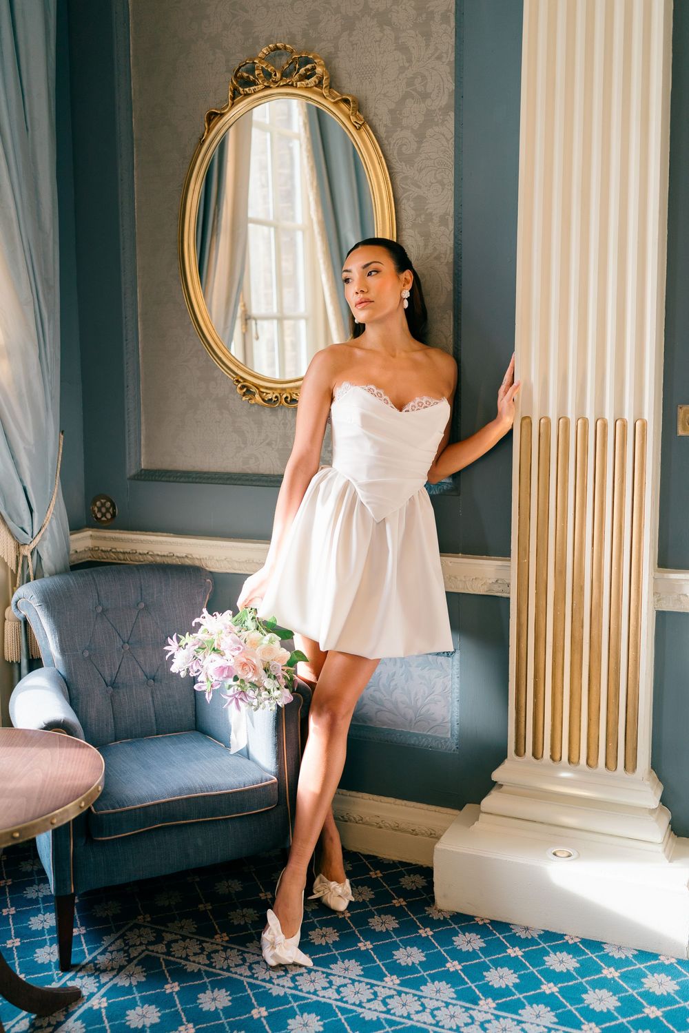 Woman in white dress poses elegantly on blue sofa in luxurious room with ornate gold mirrors and columns.