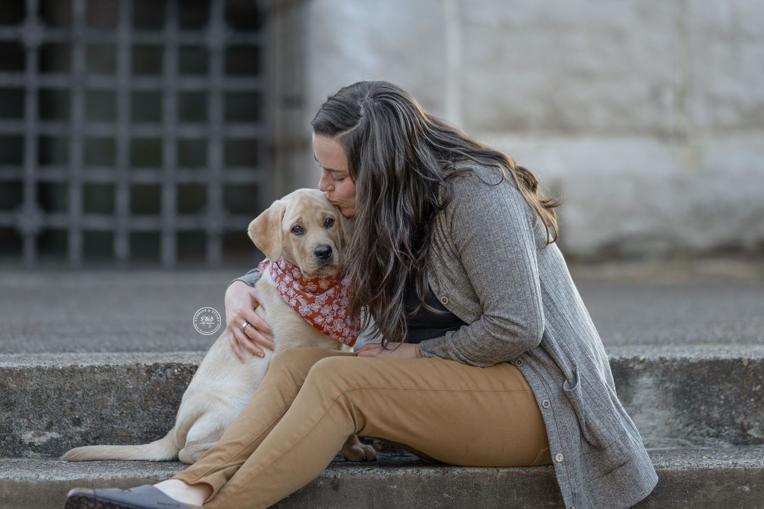 A person in a gray sweater and khaki pants sits on steps embracing a golden retriever wearing a red bandana.