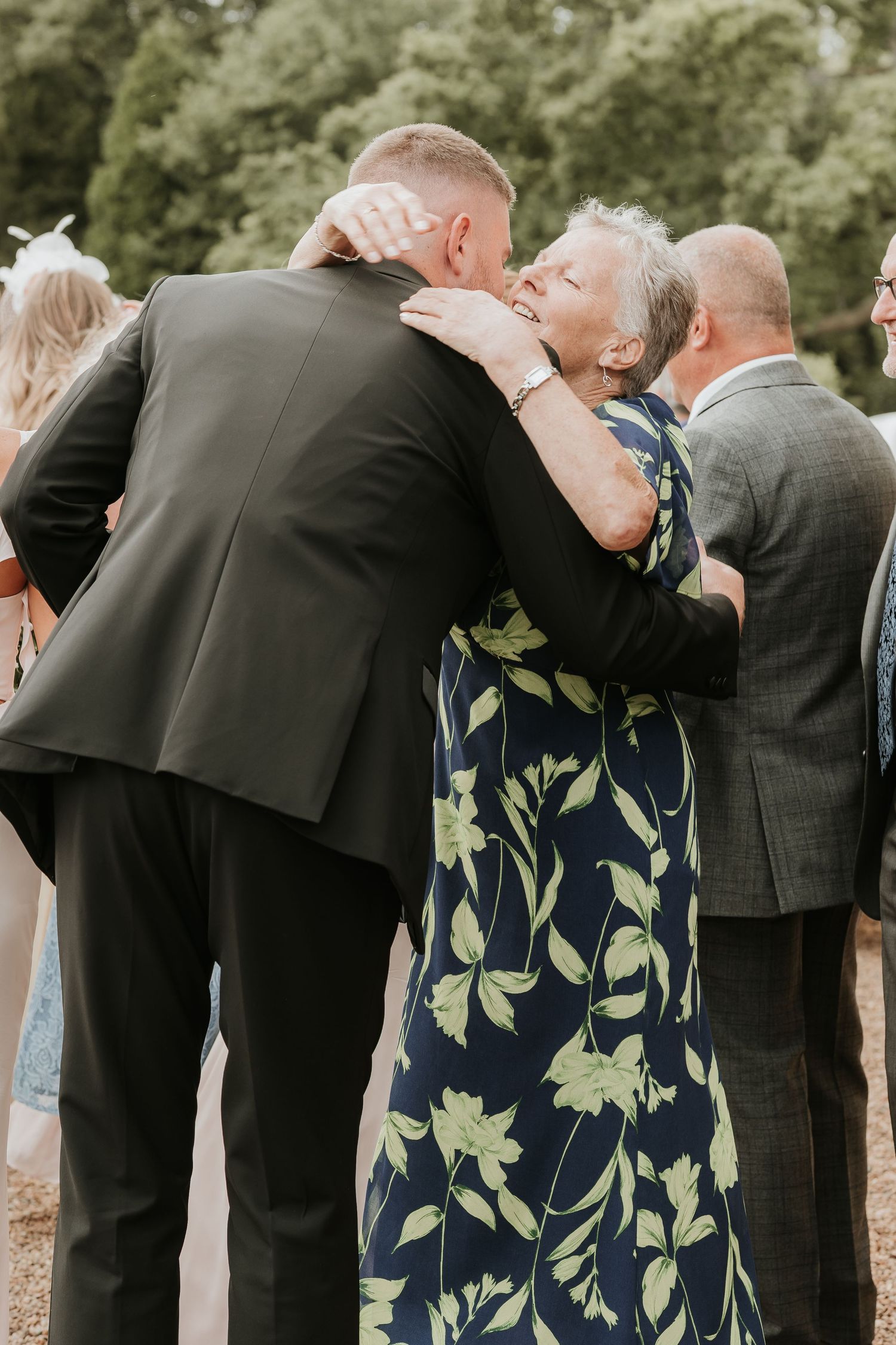Wedding guests in formal attire lean over a railing together while looking at something below during an outdoor ceremony.
