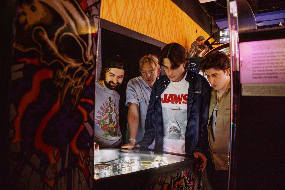 Four young men huddle around an arcade game in a dark venue, one wearing a Jaws t-shirt.