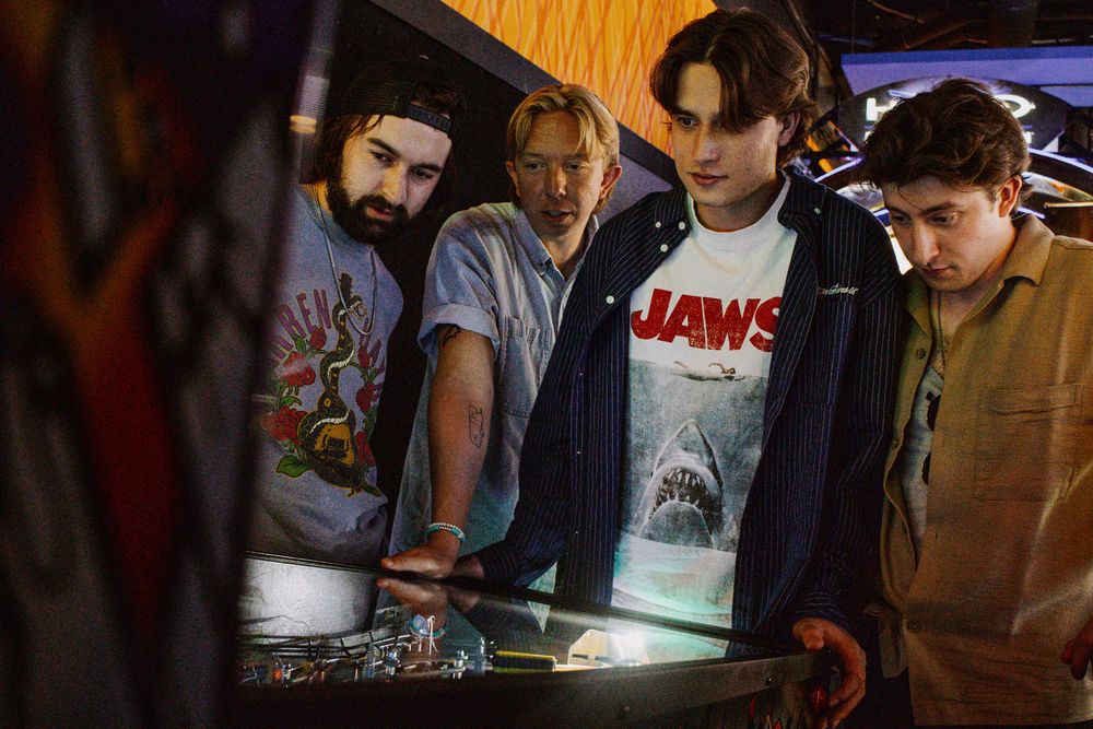 Four young men gathered around a pinball machine, one wearing a Jaws t-shirt, in a dimly lit arcade.