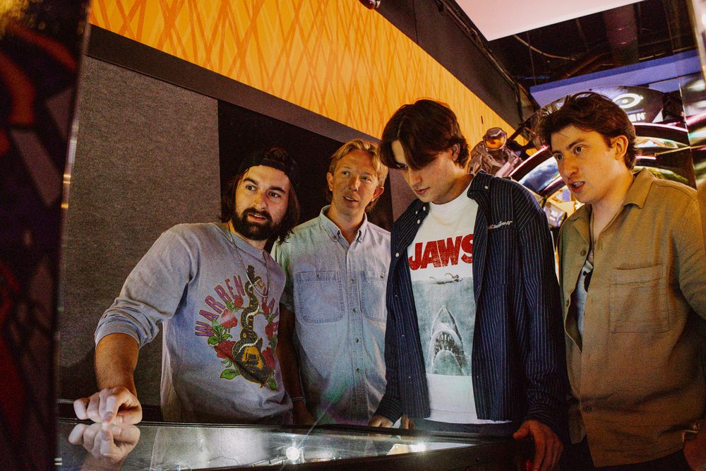 Four young men gathered around a pinball machine indoors, one wearing a Jaws t-shirt.