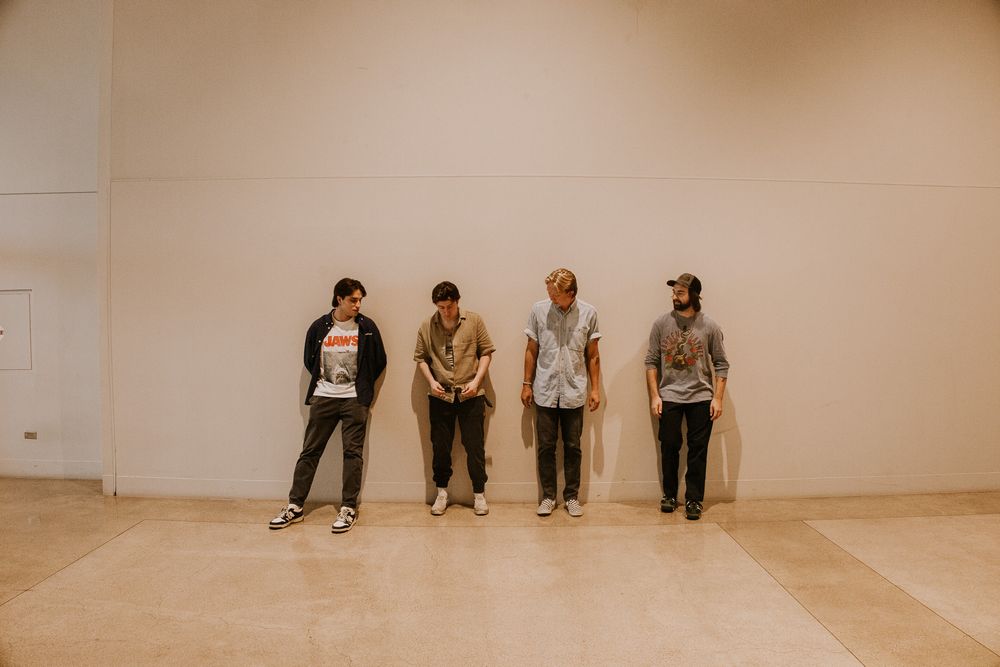 Four young men standing against a beige wall in a minimalist indoor space, wearing casual streetwear outfits.
