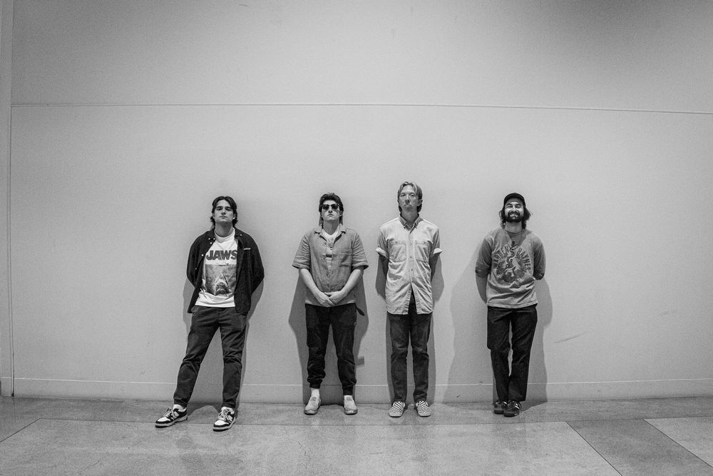 Black and white photo of four young men standing against a white wall in a casual band portrait.