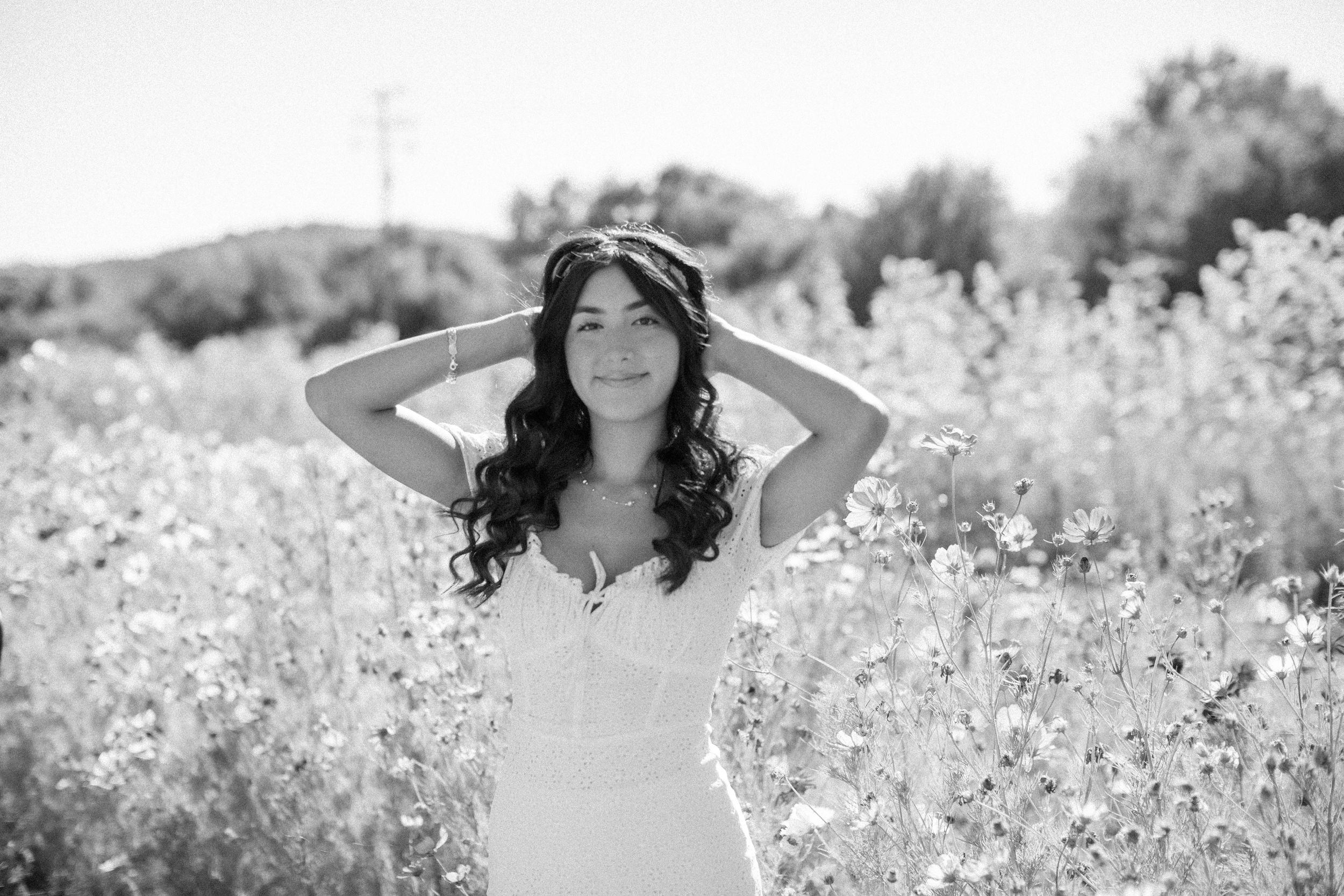 Woman in a flowing white dress poses gracefully in a blooming wildflower field on a sunny summer day.