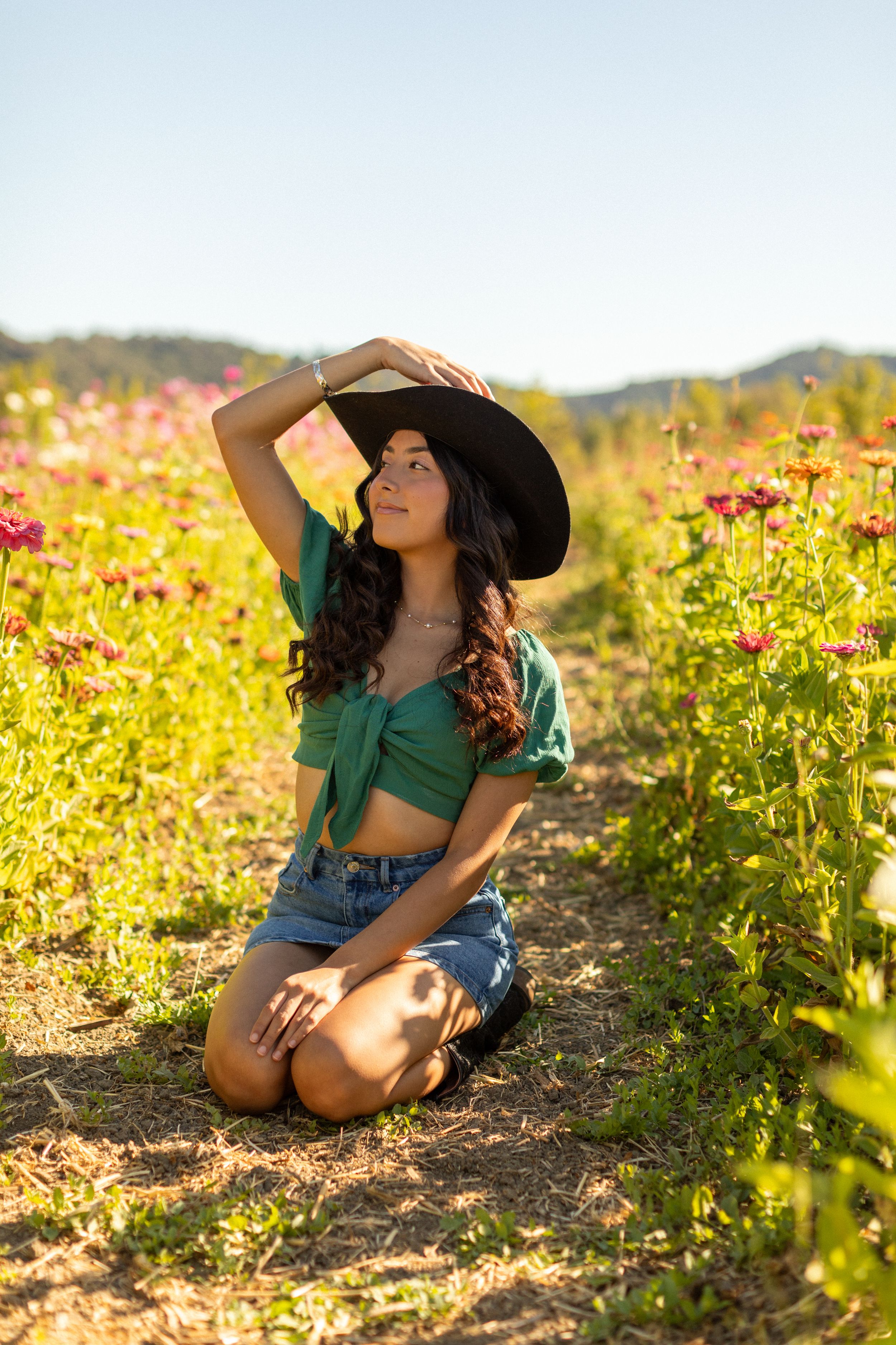 A woman in a green top and sun hat poses in a vibrant wildflower field during golden hour sunset.
