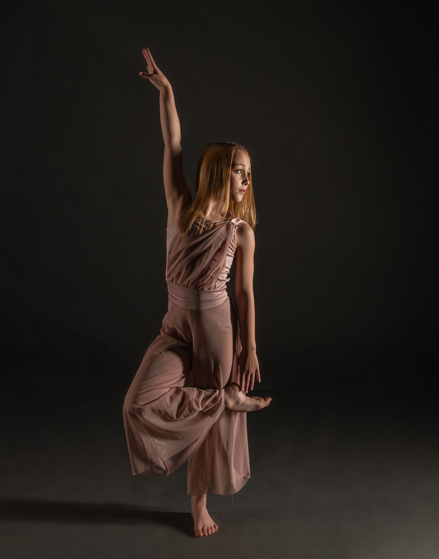 A graceful dancer in a flowing mauve dress performs an elegant leap against a dark background.