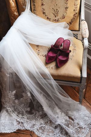 Elegant lace wedding dress detail with burgundy velvet shoes on vintage furniture.