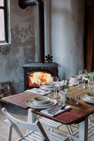 Rustic dining table set with plates and glasses near a wood-burning stove.