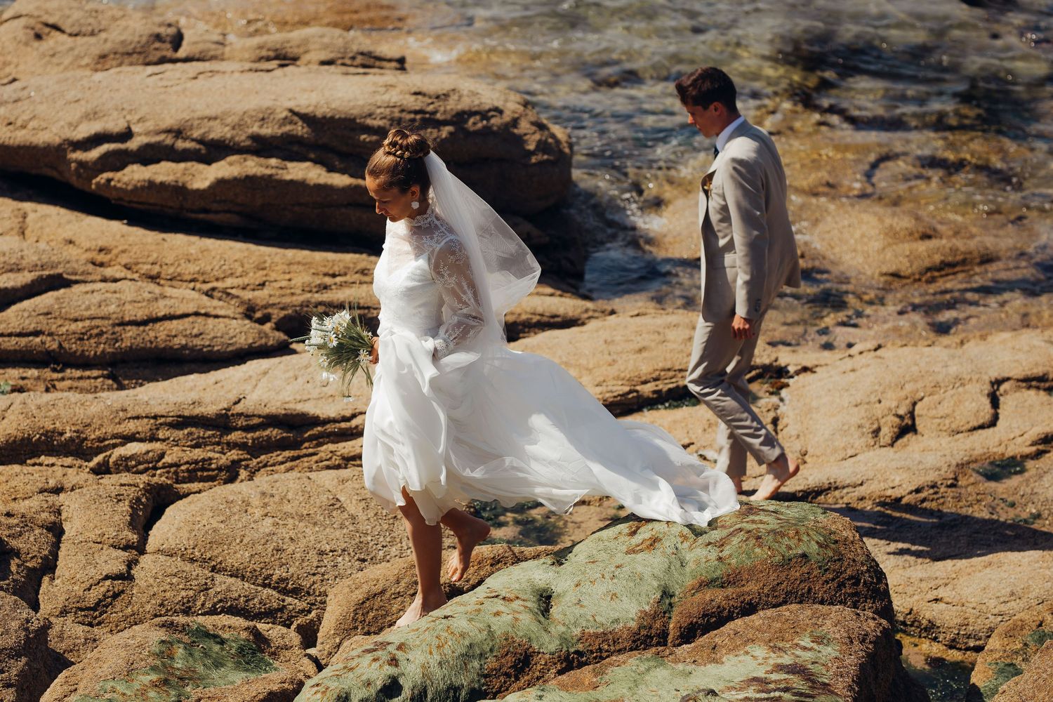 Bride in flowing white dress and groom walking on rocky coastal shoreline during wedding photoshoot.