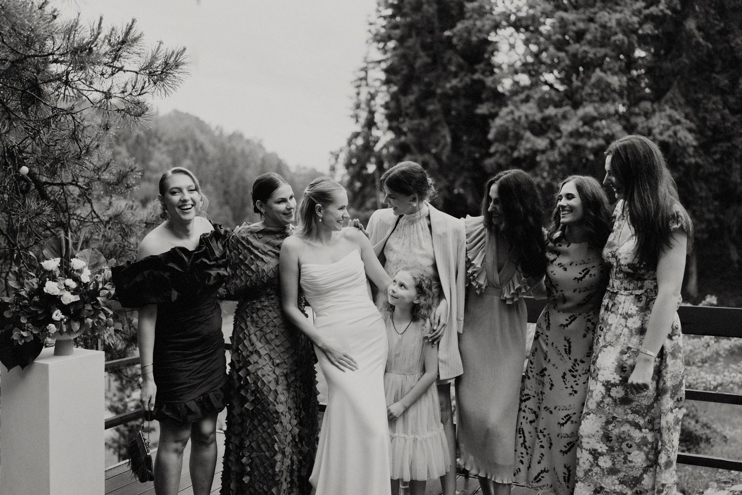 Group of people in formal attire gathering outdoors among pine trees.
