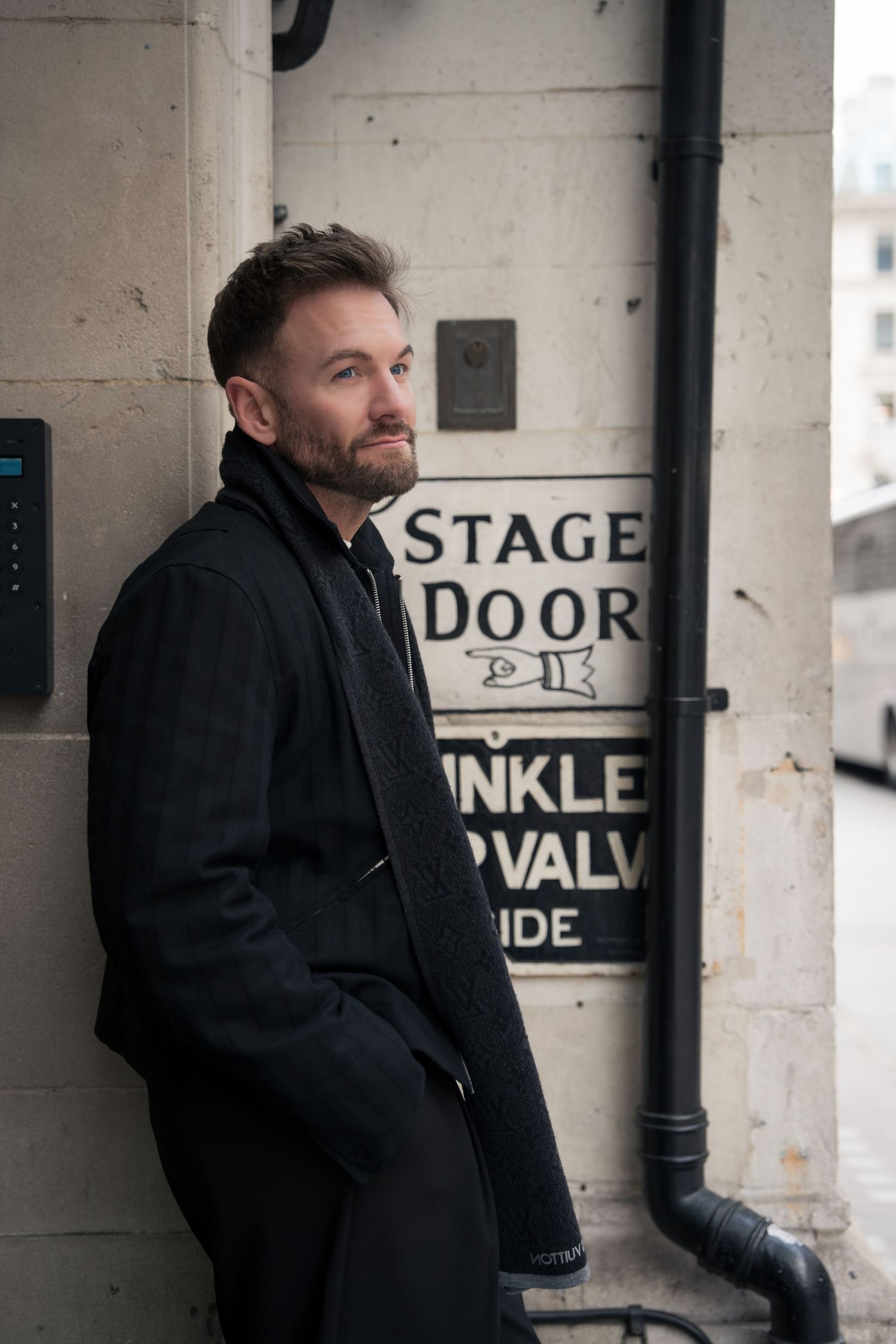 A person in a black coat stands against a concrete wall with stage door signage in an industrial setting.