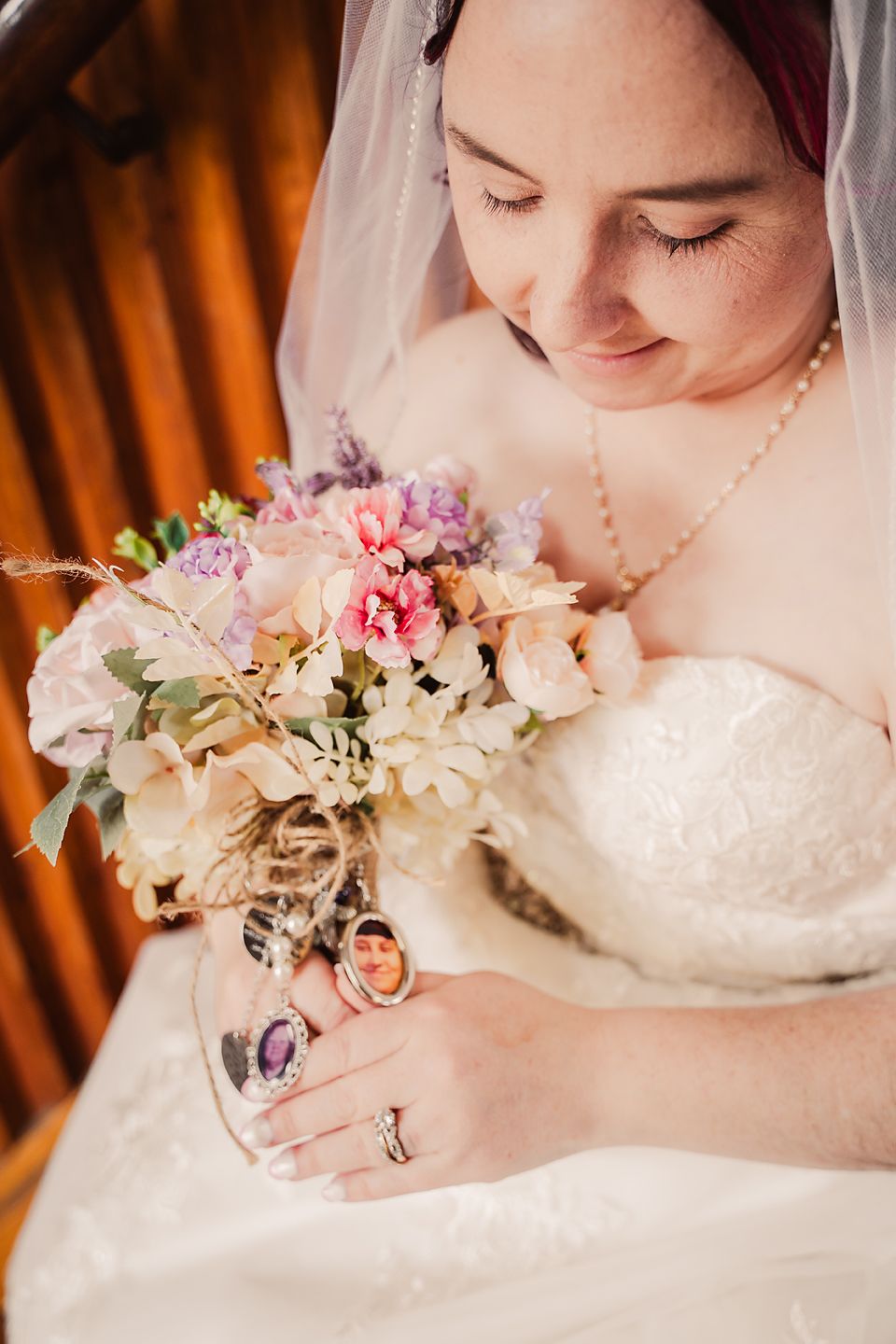 A bride in a white dress holds a delicate bouquet of pink and purple flowers while looking down with a gentle smile.