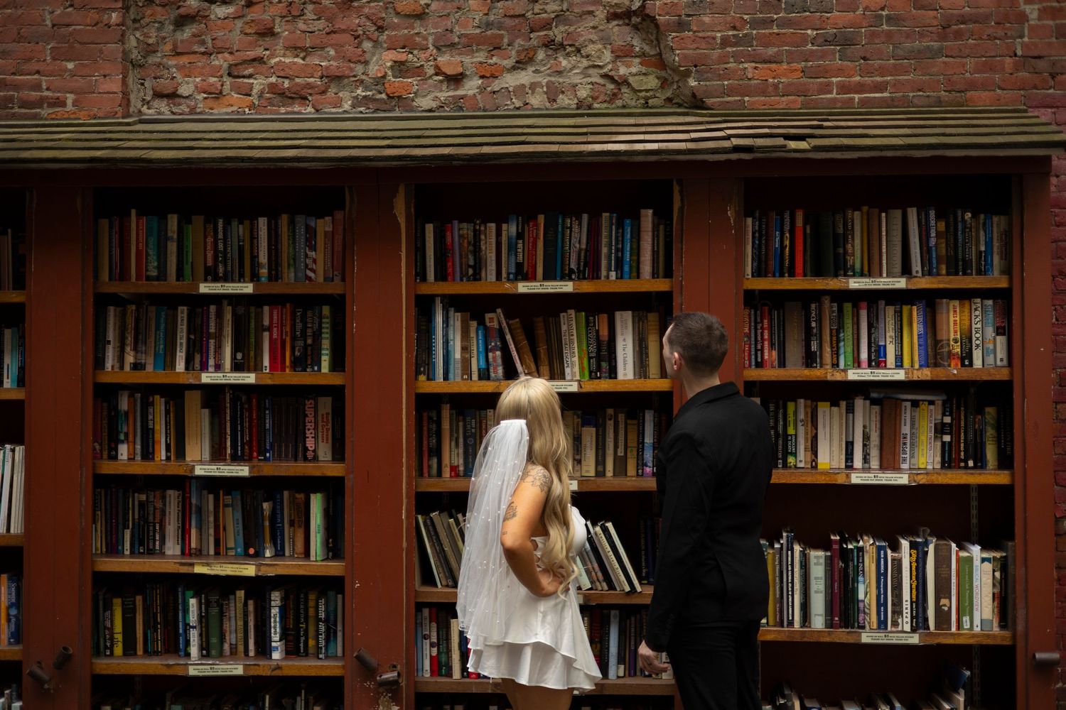 A row of library bookshelves with exposed brick walls shows students studying at wooden tables.