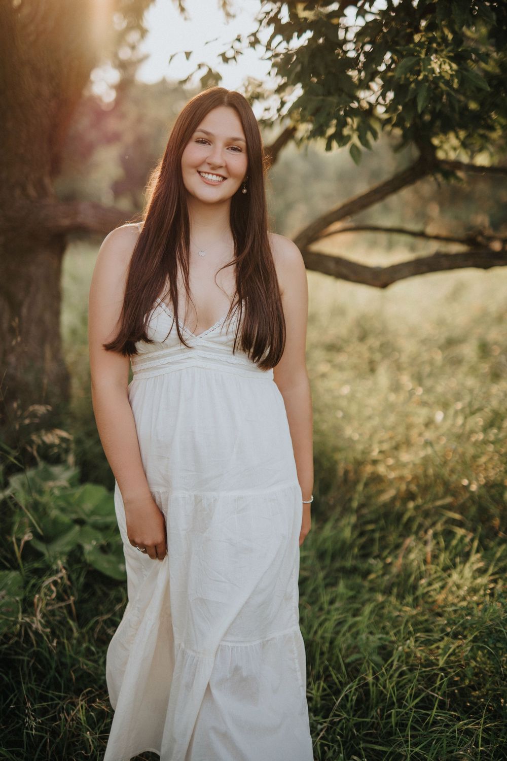 A woman in a white summer dress poses outdoors in a garden setting with warm sunlit trees in the background.