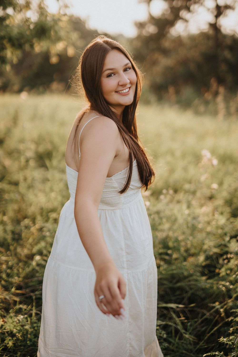 A person in a white dress poses joyfully in a sunlit meadow during golden hour.