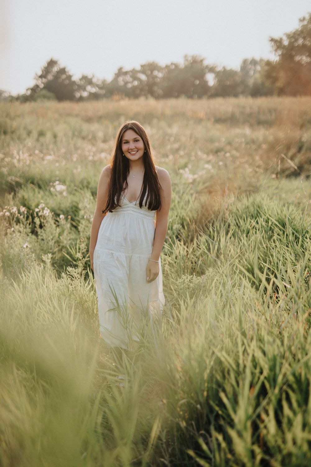 A person in a white dress stands in a sunlit meadow with tall grass and wildflowers on a summer afternoon.