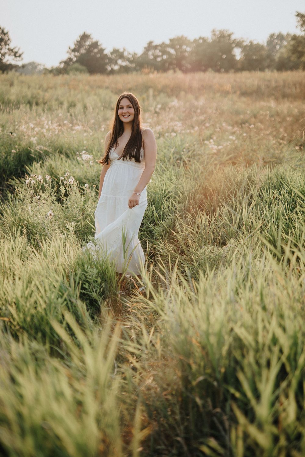 A person in a white dress walks through tall grass in a sunlit meadow during golden hour.
