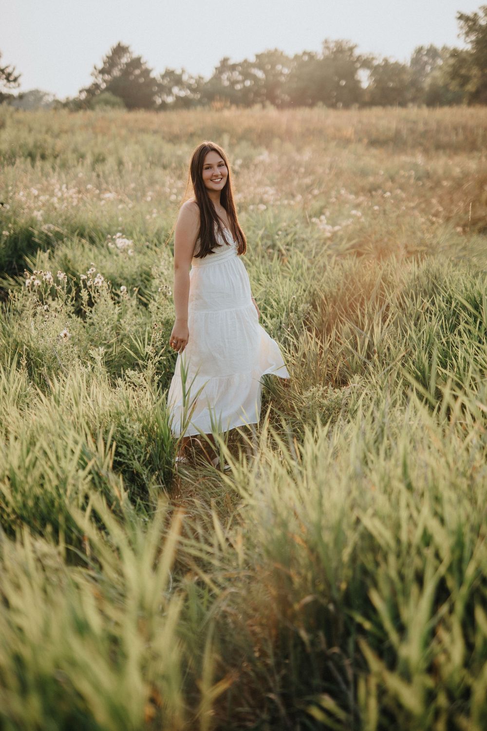 A white dress flows gracefully through tall grass in a sun-drenched field during golden hour.