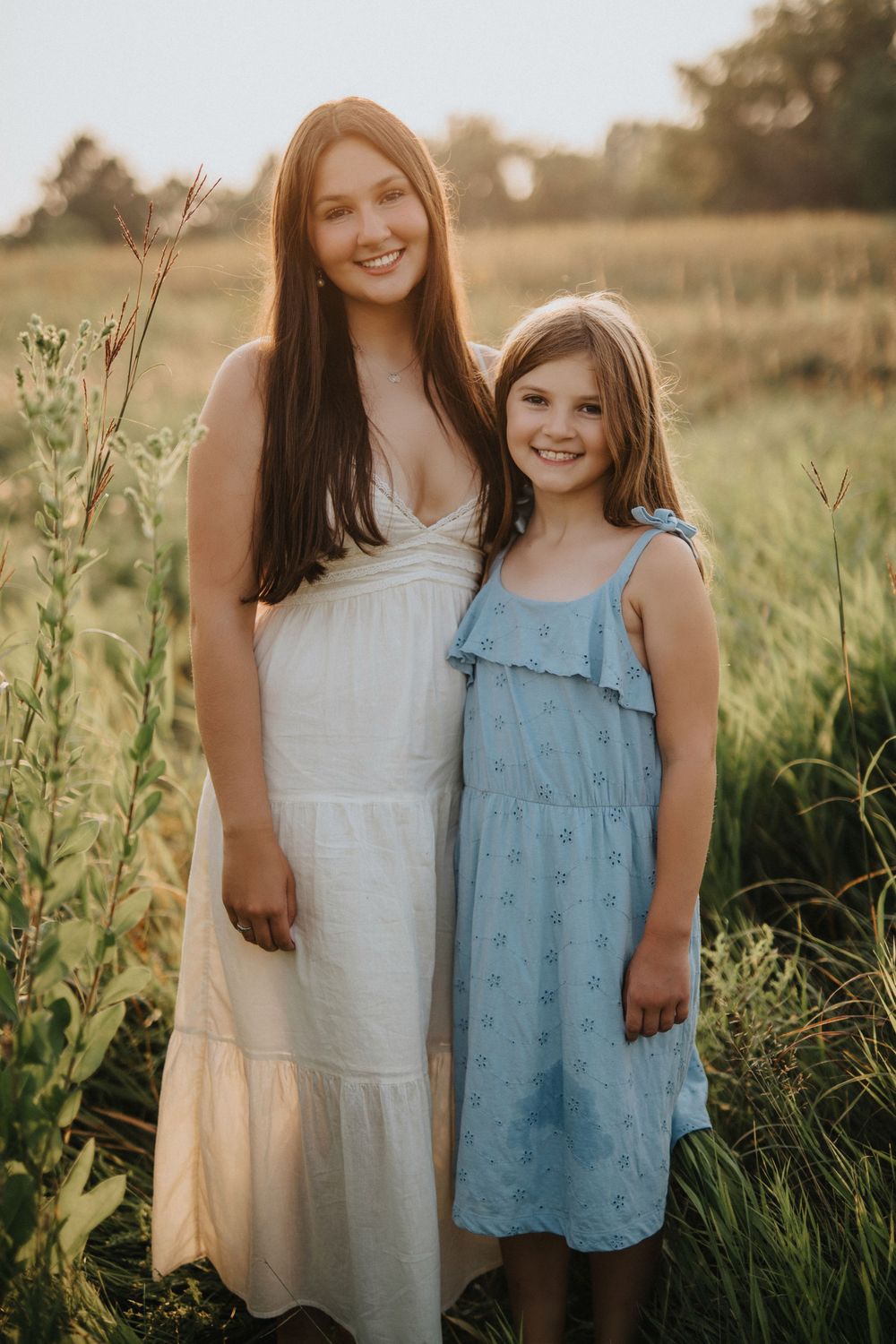 Two people in light summer dresses stand together in a golden field at sunset with tall grass and wildflowers around them.