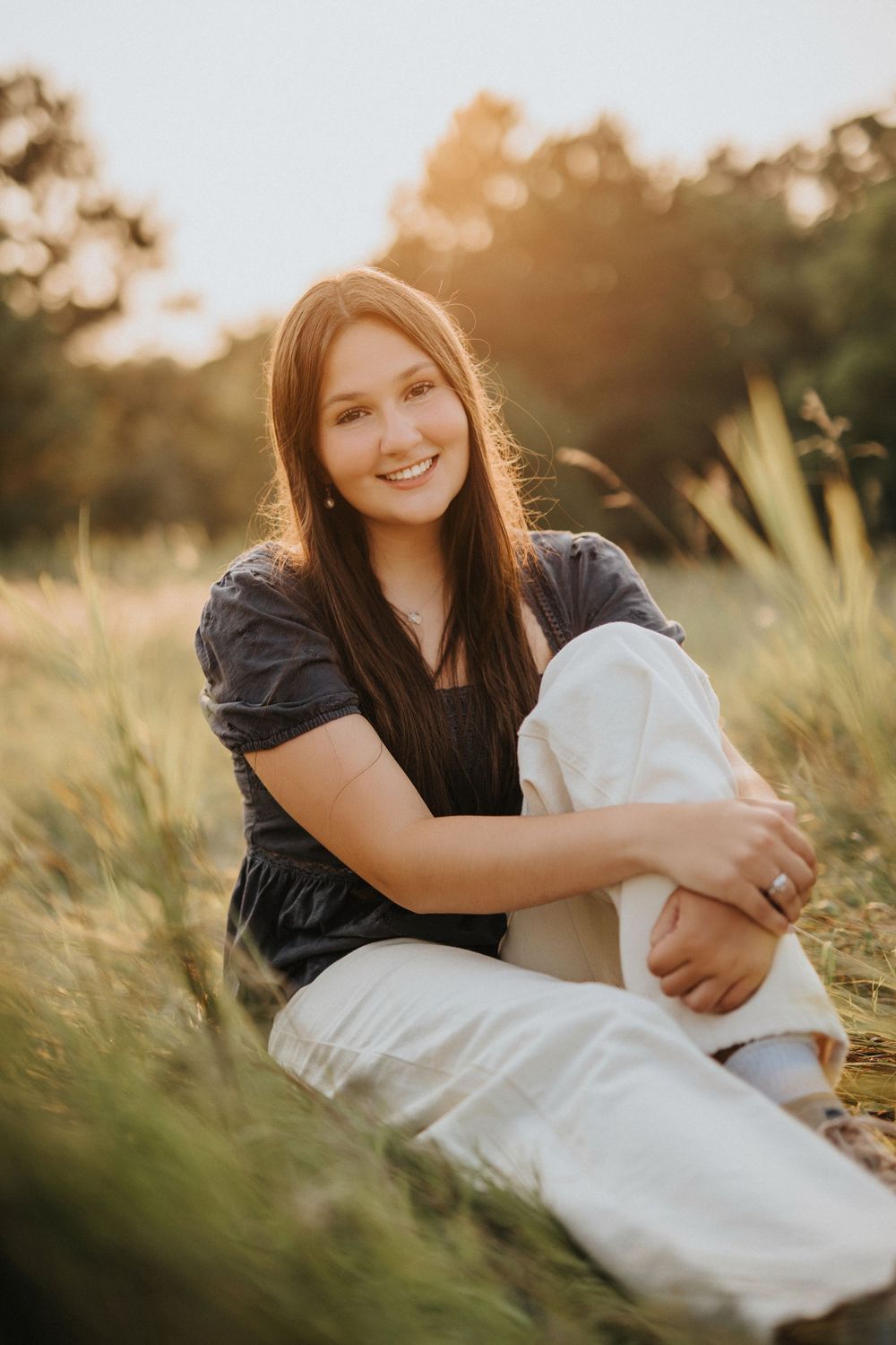 A person in a black top sits in golden sunlit grass during a warm sunset photo session.