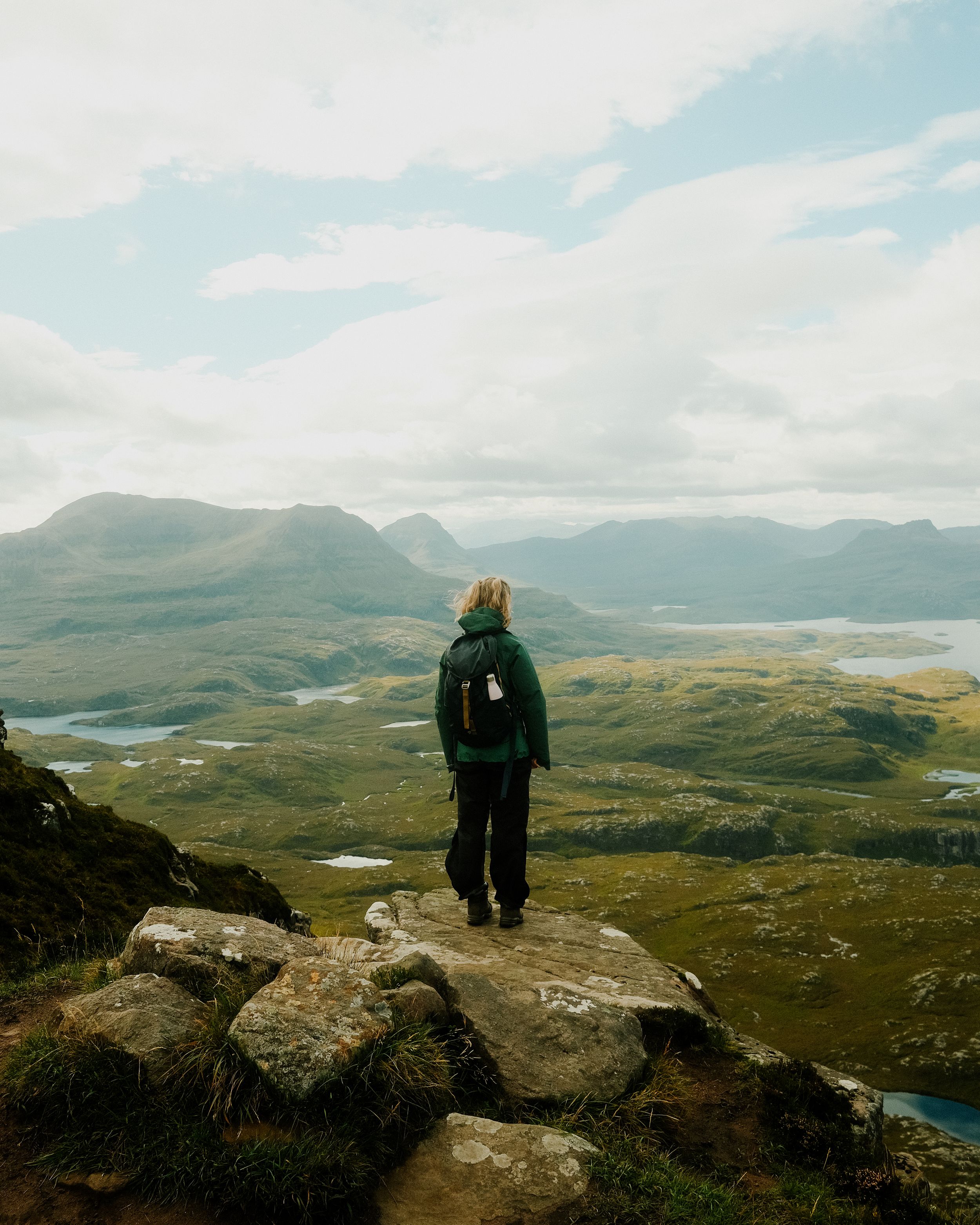 Hiker stands on rocky outcrop overlooking scenic Scottish Highlands with mountains and lakes stretching to horizon.