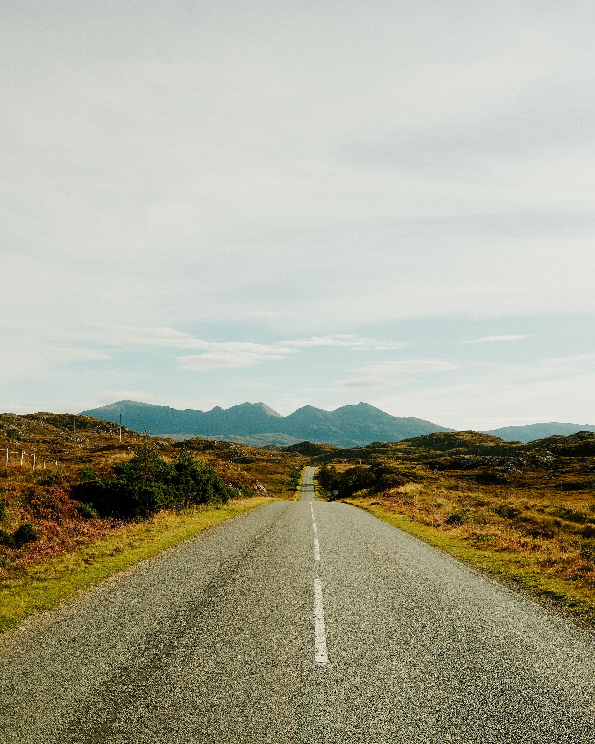 A long empty rural road stretches into the distance with mountains and rugged terrain visible on the horizon.