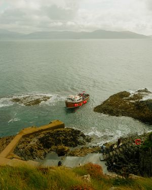 Scenic coastal view of a rugged shoreline with a red boat navigating near rocky cliffs and green hills in misty conditions.