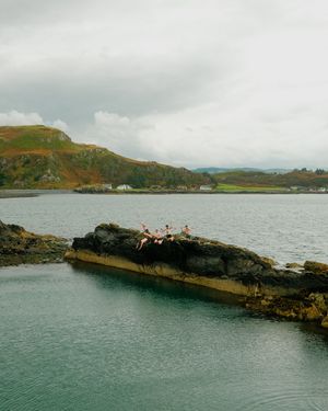 A sequence of people diving into the ocean from a rocky cliff formation on an overcast day by the coastline.