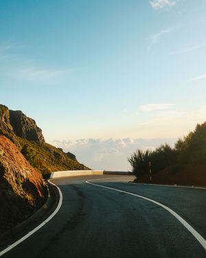 A winding mountain road curves along a coastal cliff during golden hour with dramatic ocean views in the distance.