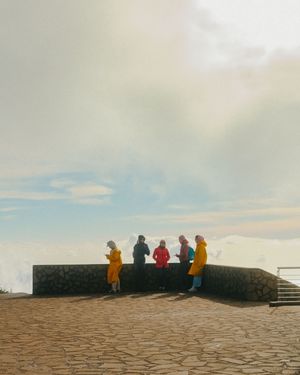 A group of people in colorful jackets sit along a stone wall overlooking a scenic vista at dusk.