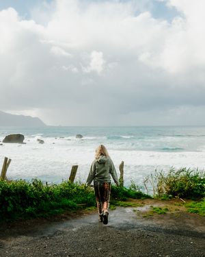 Person in green jacket walking on coastal path overlooking rocky ocean shore and cloudy sky.