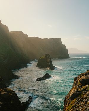 Dramatic coastal landscape with rugged cliffs and turquoise waves crashing against rocky shores at sunset.