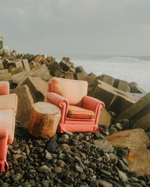 Pink vintage sofa positioned dramatically on rocky beach against dark stormy ocean waves and boulders.