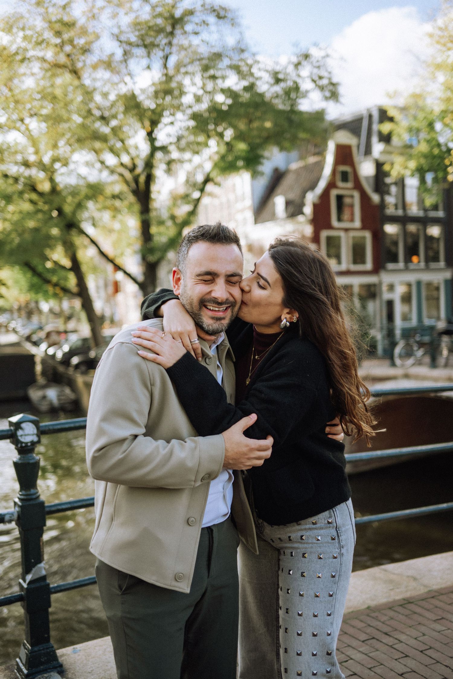 A couple shares a romantic moment together while laughing on a bridge in Amsterdam during autumn after secret proposal.