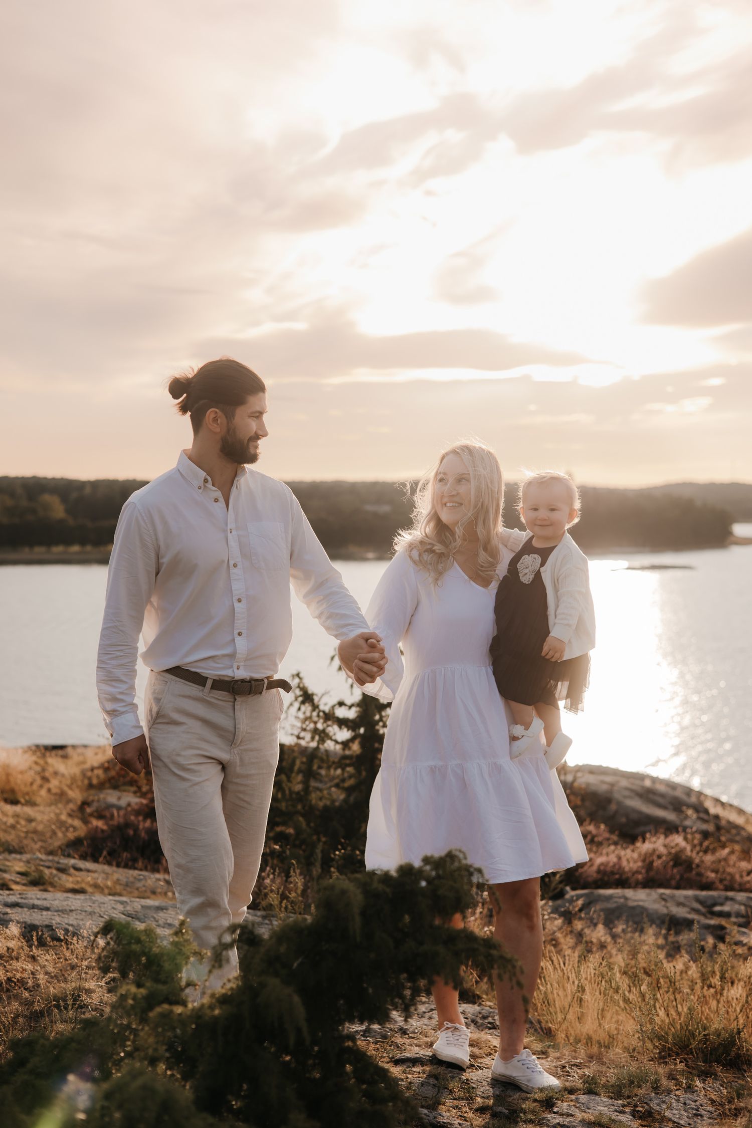A family in white outfits holds hands while standing together by a serene lakeside at sunset.