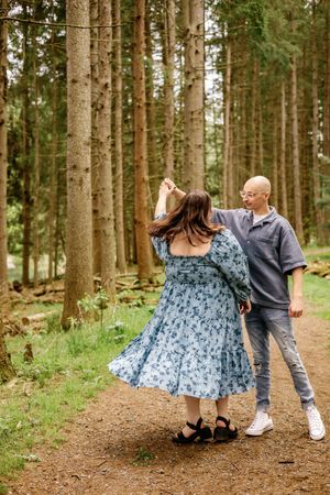 Playful couple dancing on forest path surrounded by tall pine trees.