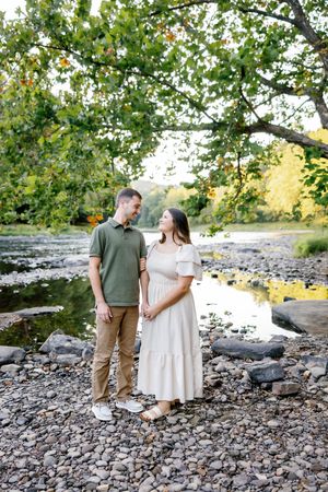 A couple stands together on a rocky riverbank surrounded by trees and natural scenery.