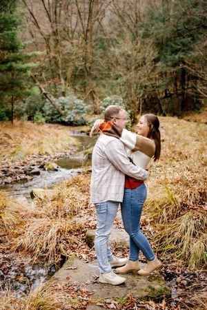 A couple shares a tender moment by a stream in an autumn forest setting.