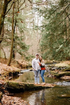 A couple stands together beside a peaceful forest stream during autumn.