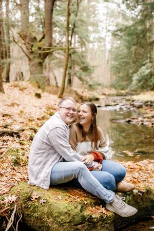Two people share a joyful moment while sitting on a log by a creek in the woods.