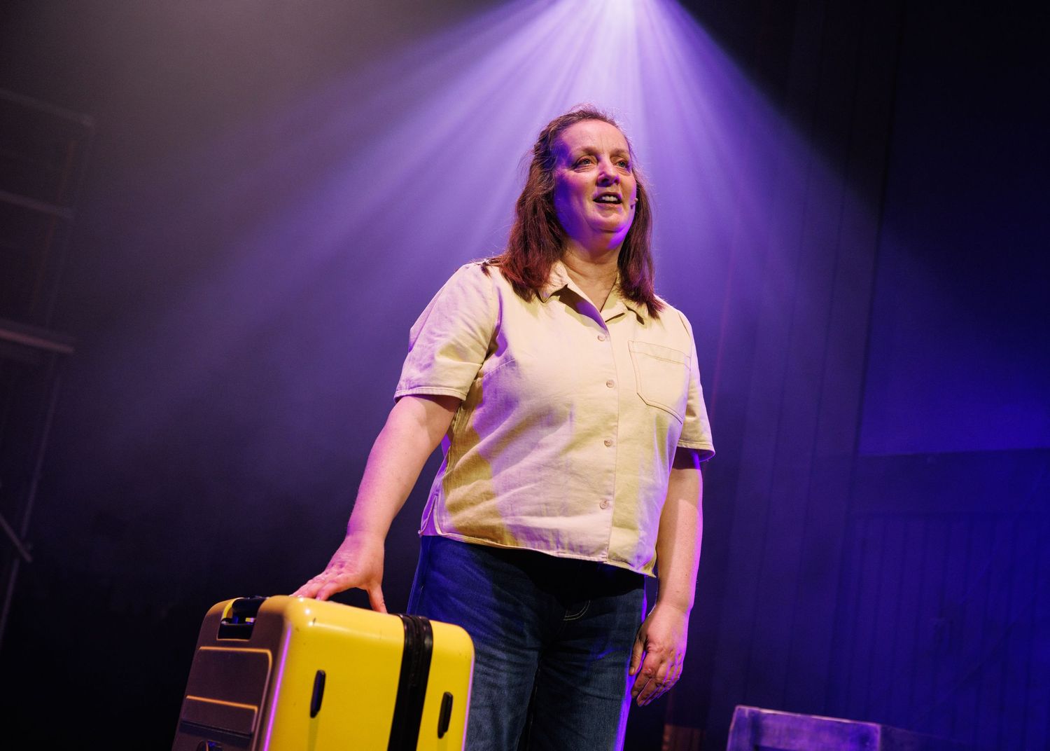 Performer on stage sits on yellow chair under dramatic purple lighting during theatrical production.