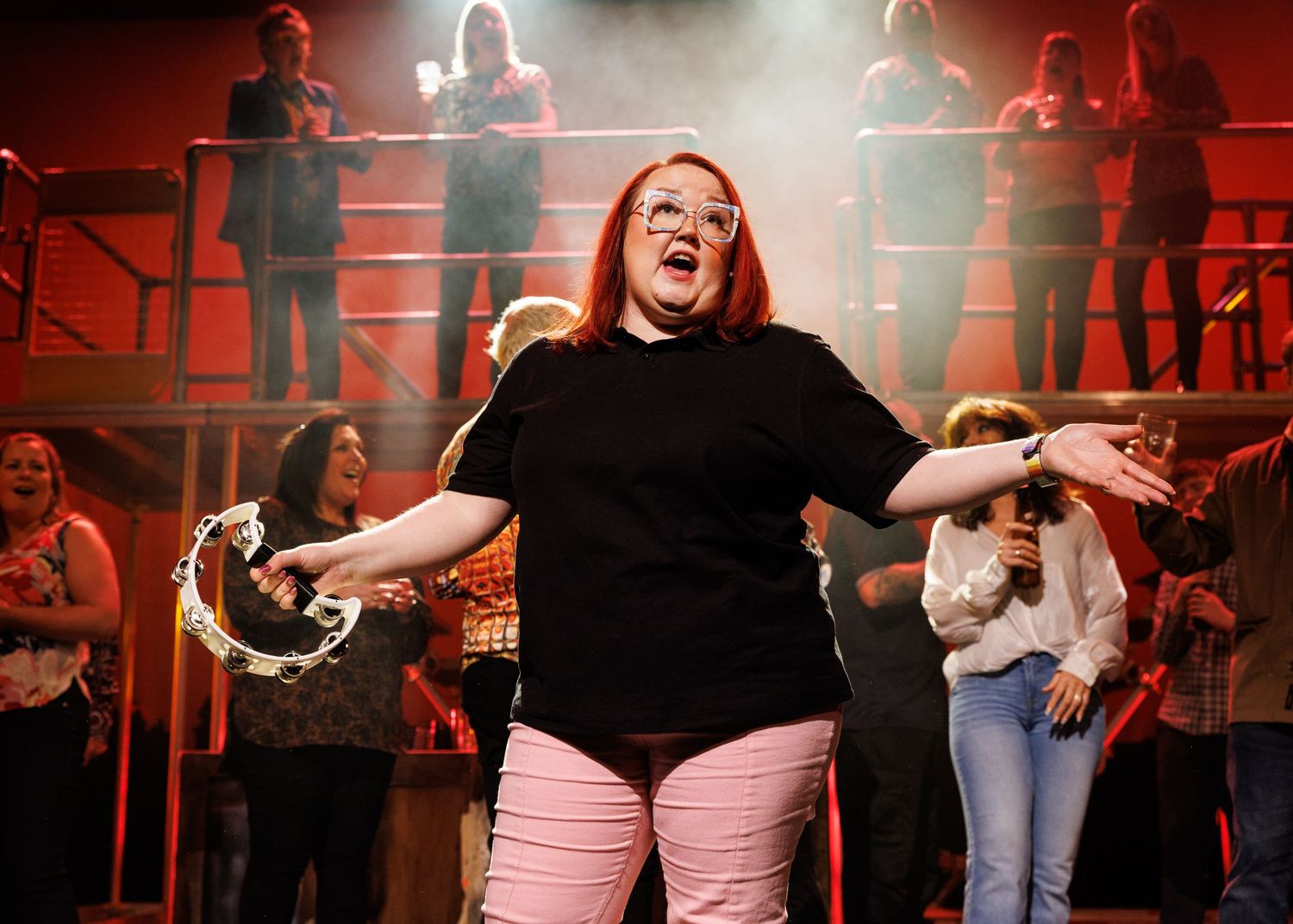 A performer in a black shirt and pink pants sings passionately on stage with backup performers on risers behind them.
