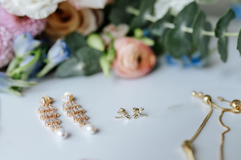 Delicate gold bridal jewelry and accessories arranged with soft pink and white flowers on a light surface.