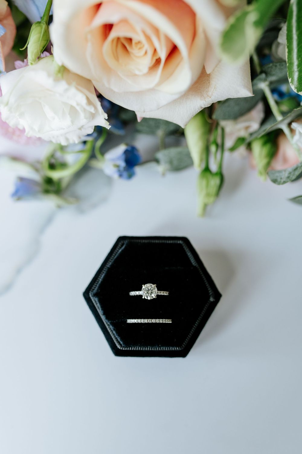 Three elegant engagement rings displayed in black hexagonal boxes with blush roses in the background.