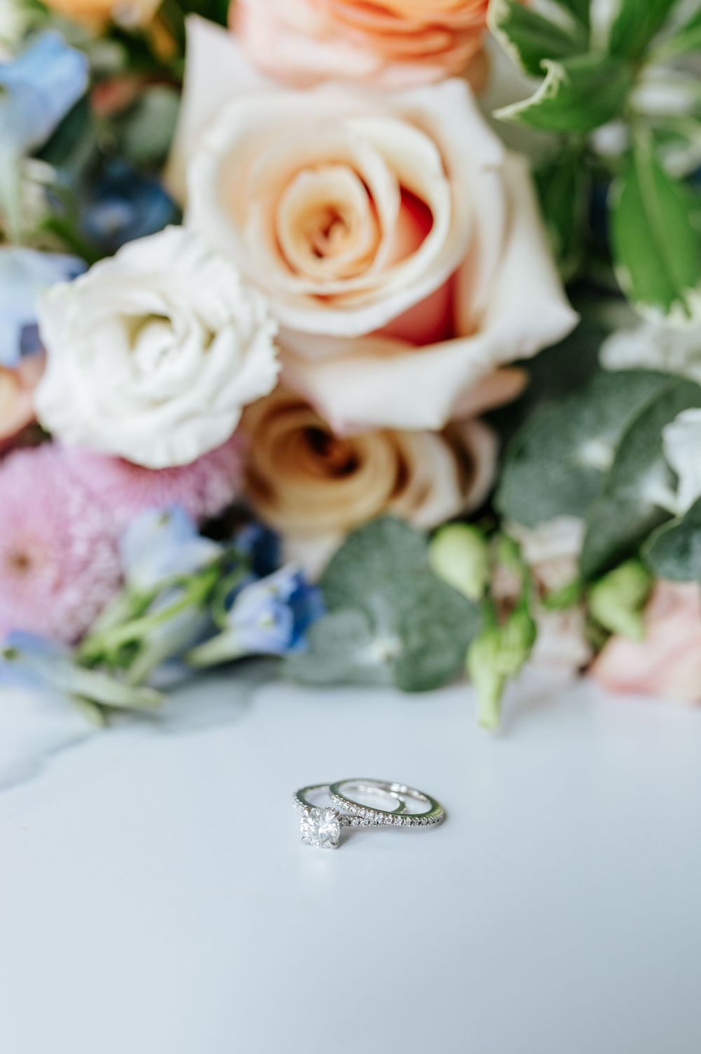 Delicate diamond engagement ring nestled among soft peach roses and white floral arrangements.