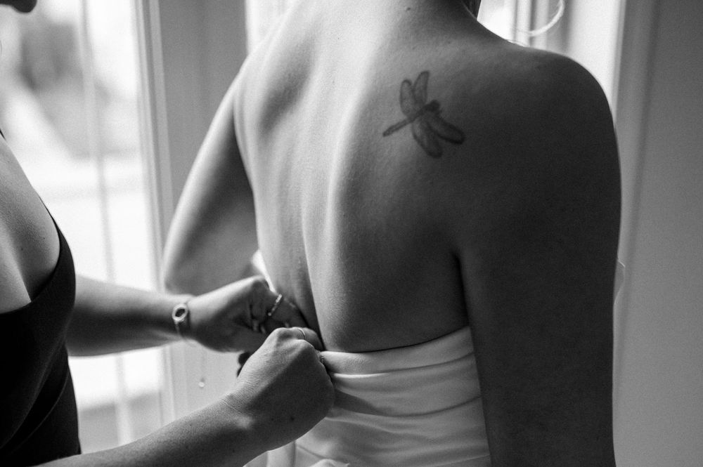 Black and white photo of someone getting dressed in white garment, showing small dragonfly tattoo on upper back.