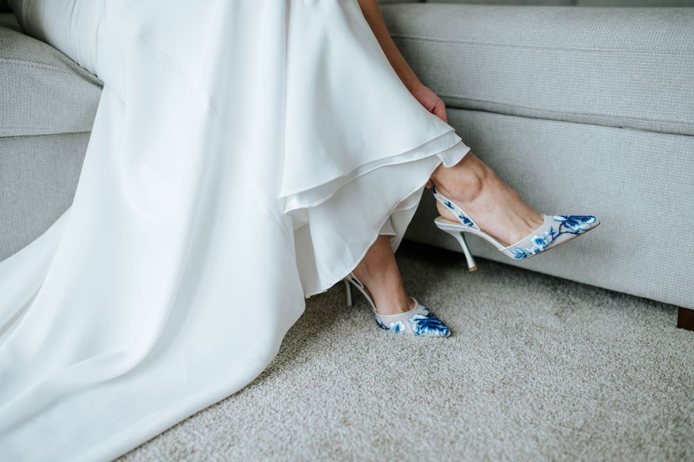 Close-up details of a bride's white dress and blue shoes while standing indoors.