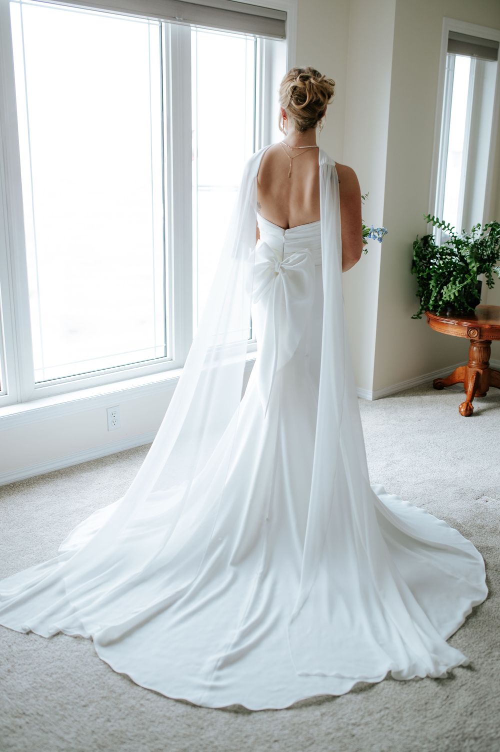 A flowing white wedding dress with a long train is displayed in a bright room with large windows.